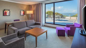 Habitación de hotel con sofá, sillas, una mesita de centro, escritorio y un ventanal con vistas al mar.