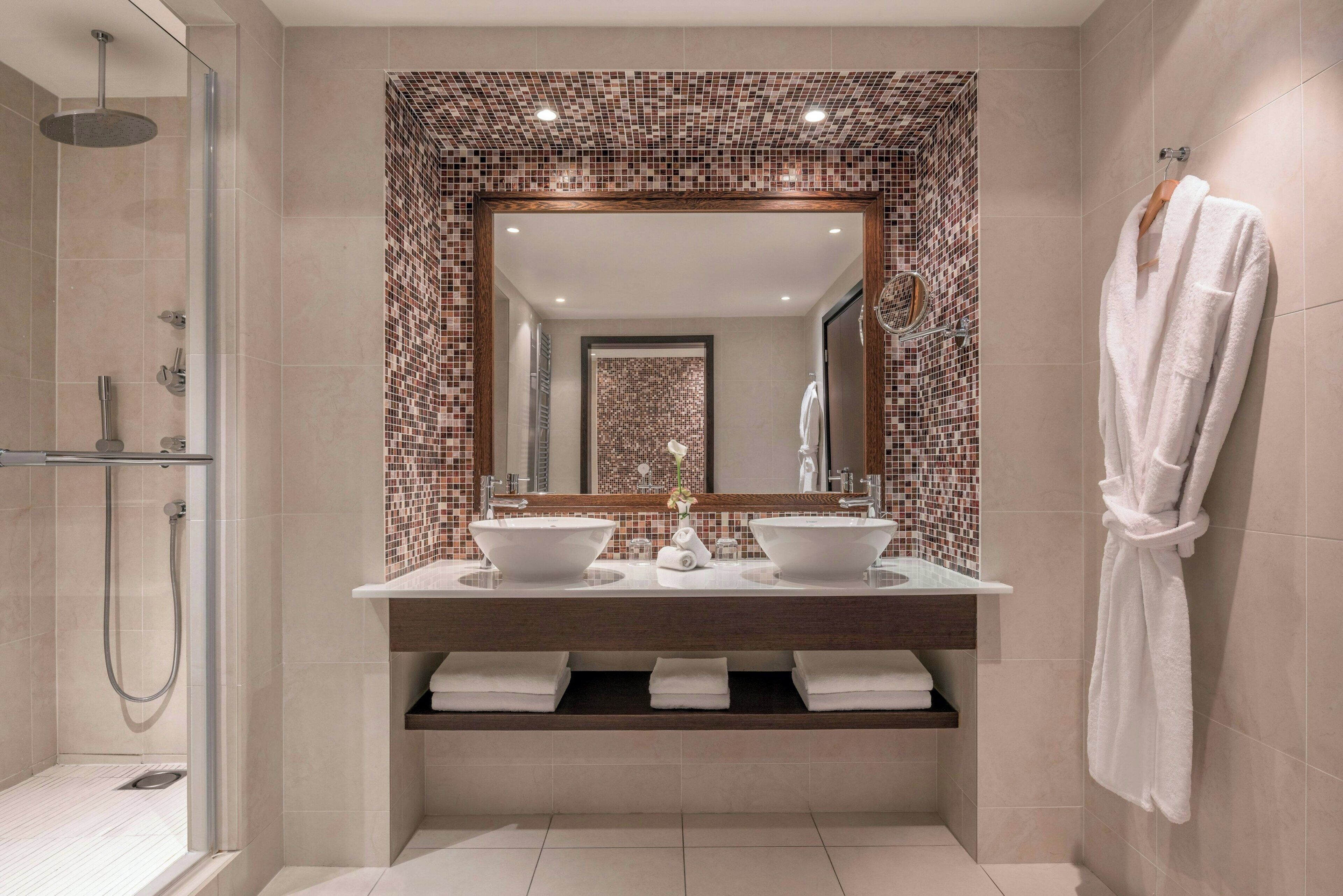 Une salle de bain moderne dotée d’une douche à paroi de verre, de deux vasques et d’un grand miroir orné d’une bordure en mosaïque.