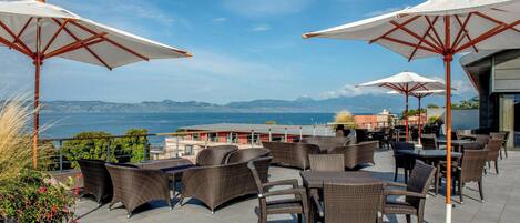 An outdoor seating area with wicker furniture and umbrellas, overlooking a body of water and distant mountains.