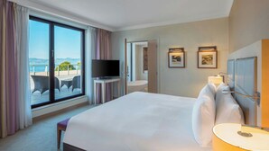 A hotel room with a large bed, a television on a stand, a bathroom with a shower, and a view of the sea. - Hotel Evian-les-Bains by Leonardo Hotels (Evian-les-Bains)
