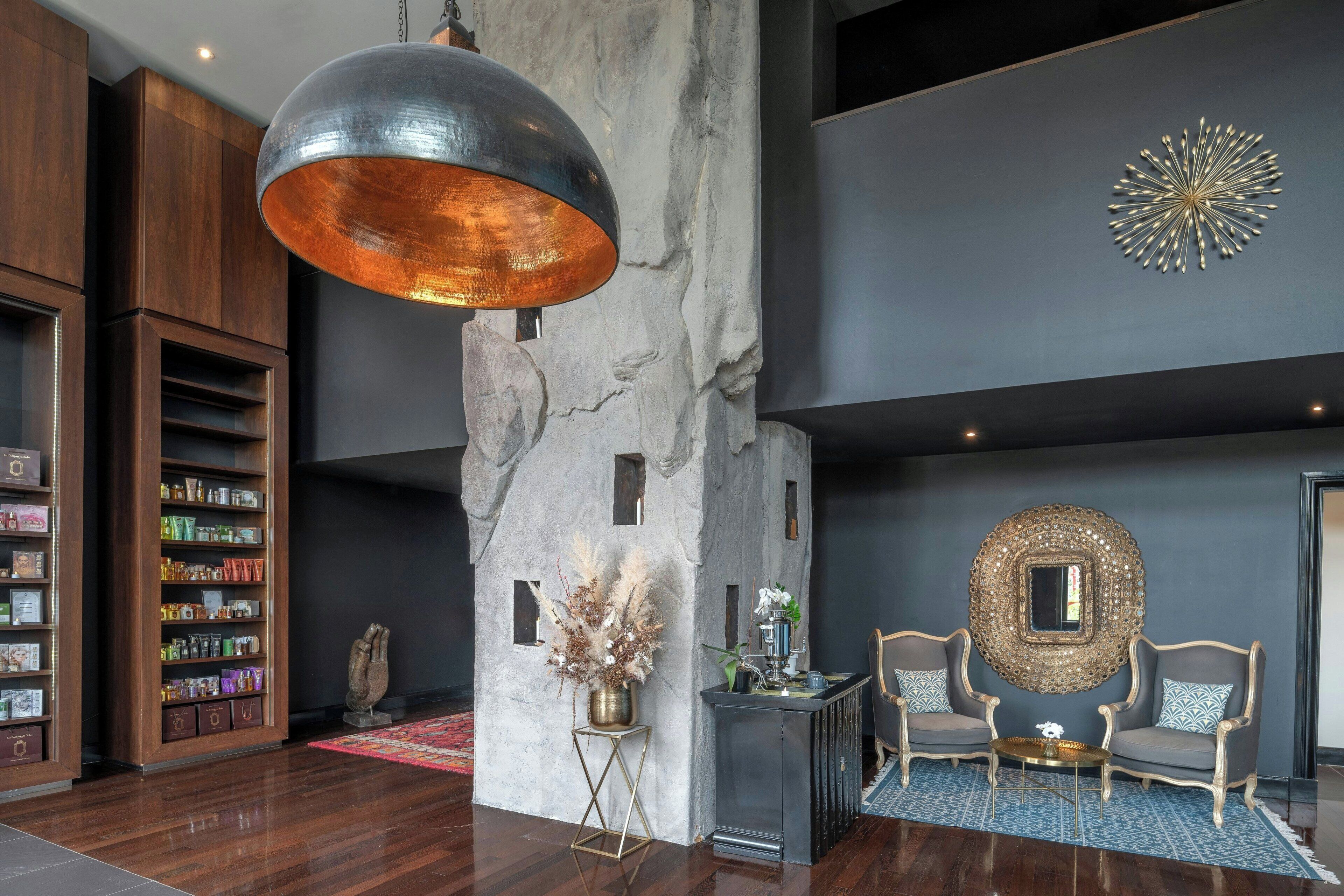 A modern lobby with a large hanging light fixture, a stone column, a decorative mirror, and a seating area with two armchairs.