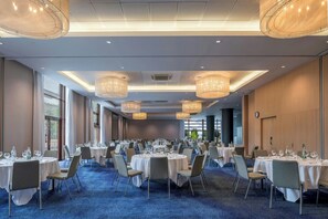 Een ruime balzaal met ronde tafels, stoelen en hangende kroonluchters.