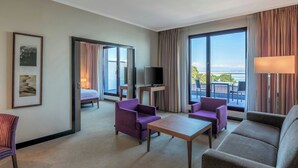 A hotel room with a balcony, a TV, purple and grey furniture, and a large window with curtains. - Hotel Evian-les-Bains by Leonardo Hotels (Evian-les-Bains)