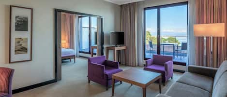 A hotel room with a balcony, a TV, purple and grey furniture, and a large window with curtains.