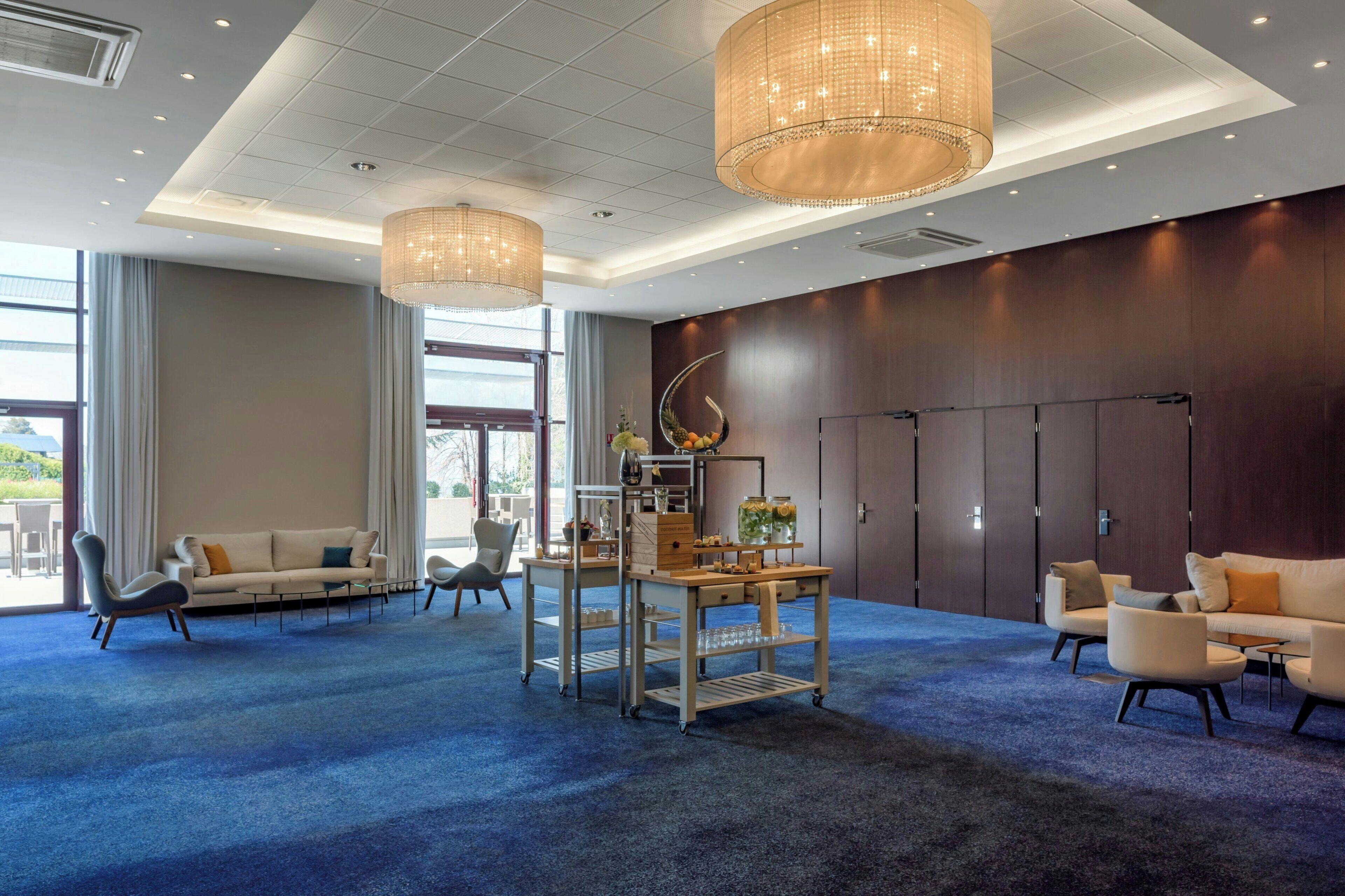 Een hotellobby met een blauw tapijt, zithoeken, een receptiebalie en een groot raam.