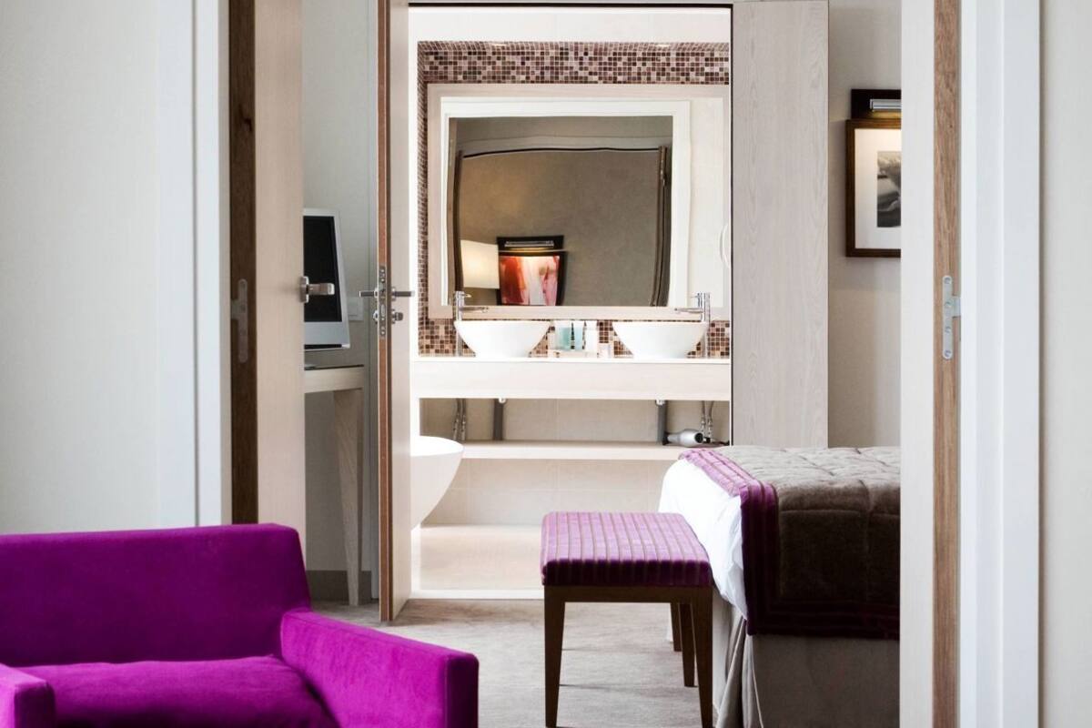 Une chambre d’hôtel avec un fauteuil violet, un lit, une salle de bain dotée d’un grand miroir et un banc en bois.