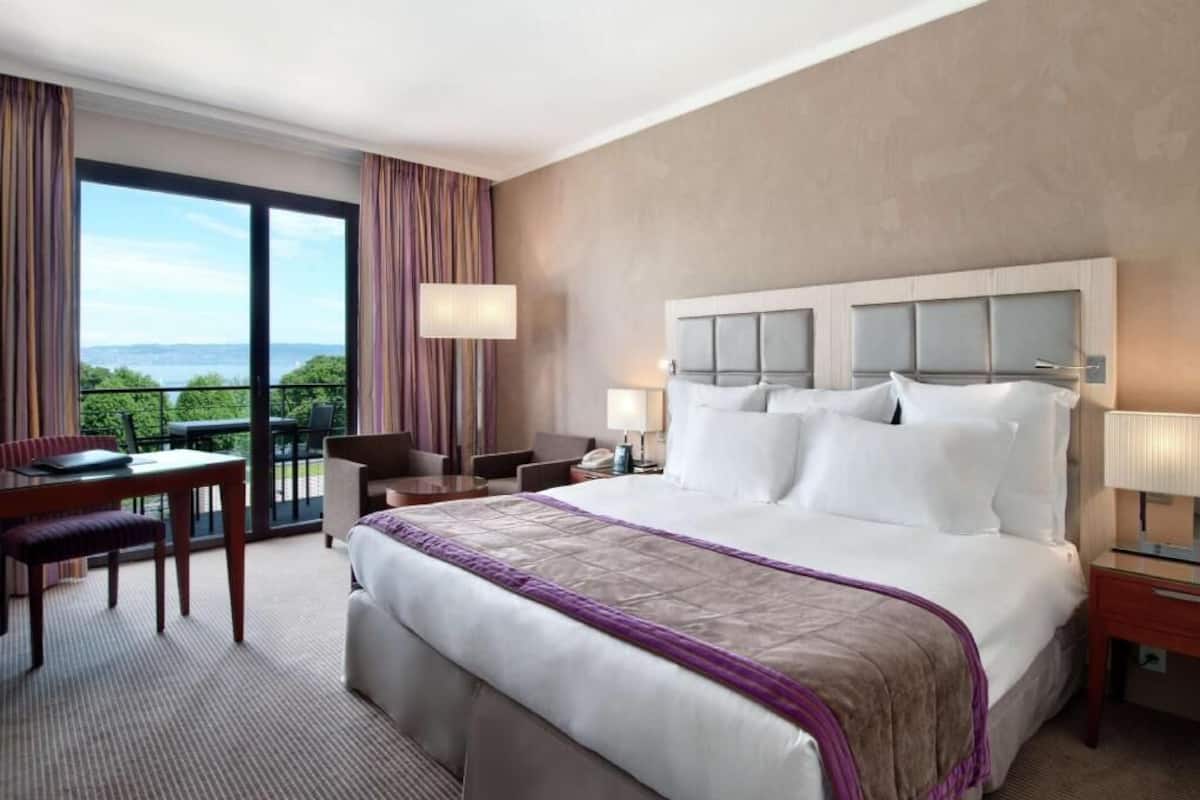 Une chambre d’hôtel avec un grand lit, un bureau, une chaise, une lampe et une vue sur la verdure par la fenêtre.