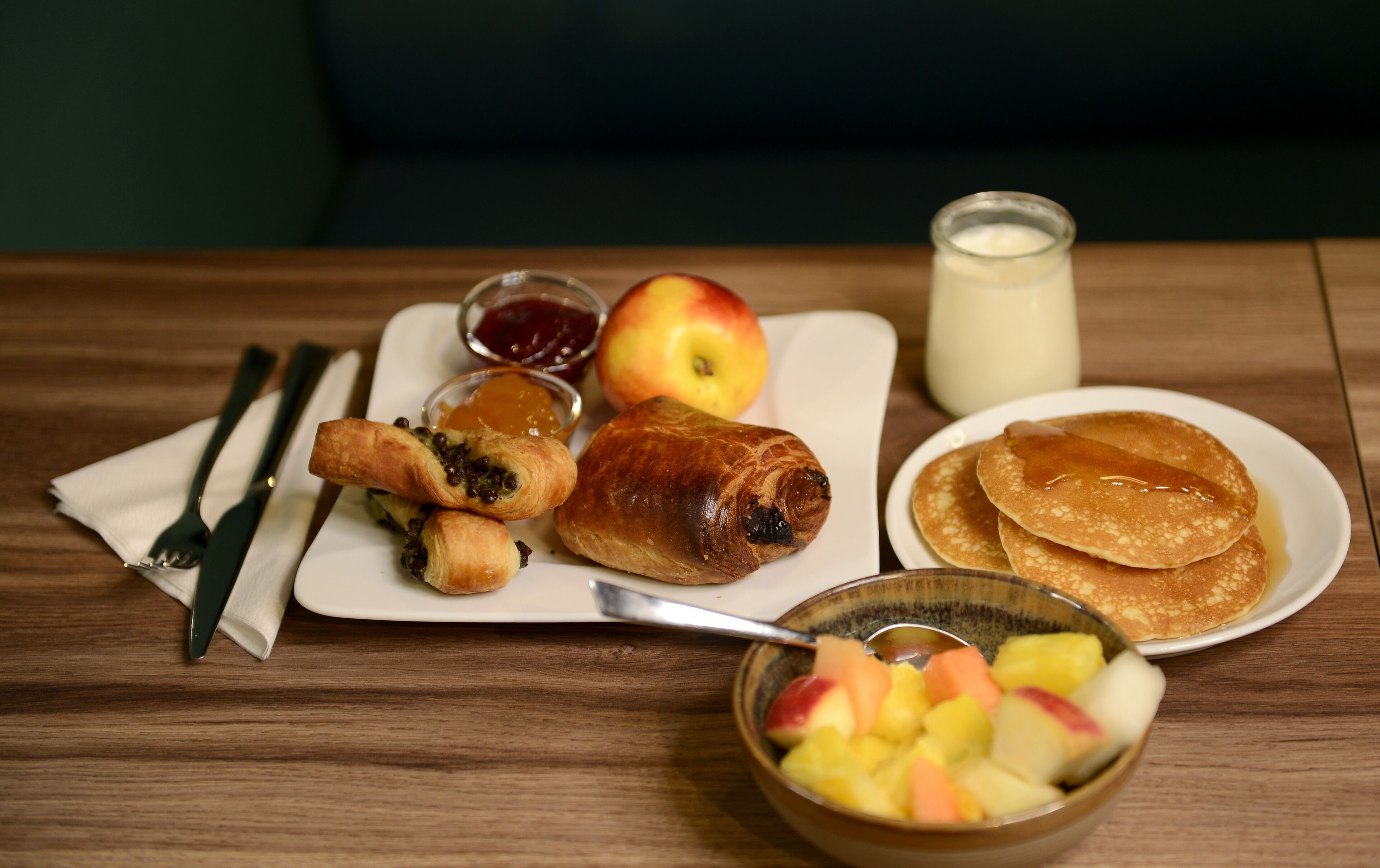 Un plato de desayuno con tortitas, un croissant, una manzana y un bol de fruta.