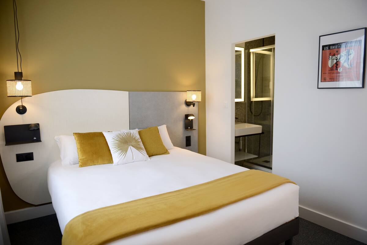 Habitación de hotel con una cama grande, dos lámparas de pared, un baño con espejo y lavamanos, y un cartel enmarcado en la pared.