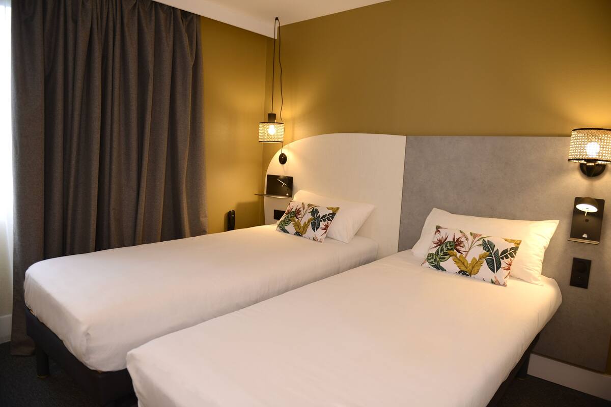 Una habitación de hotel con dos camas individuales, un ventanal con cortinas y una lámpara fijada a la pared.