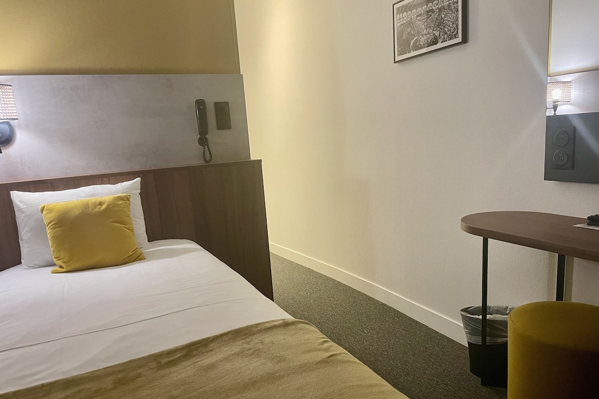 Habitación de hotel con cama, mesita de noche, silla, televisor montado en la pared y un cesto de basura.