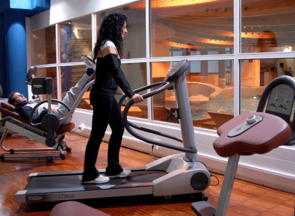 A person using a treadmill in a gym with large windows and a visible pool area in the background.