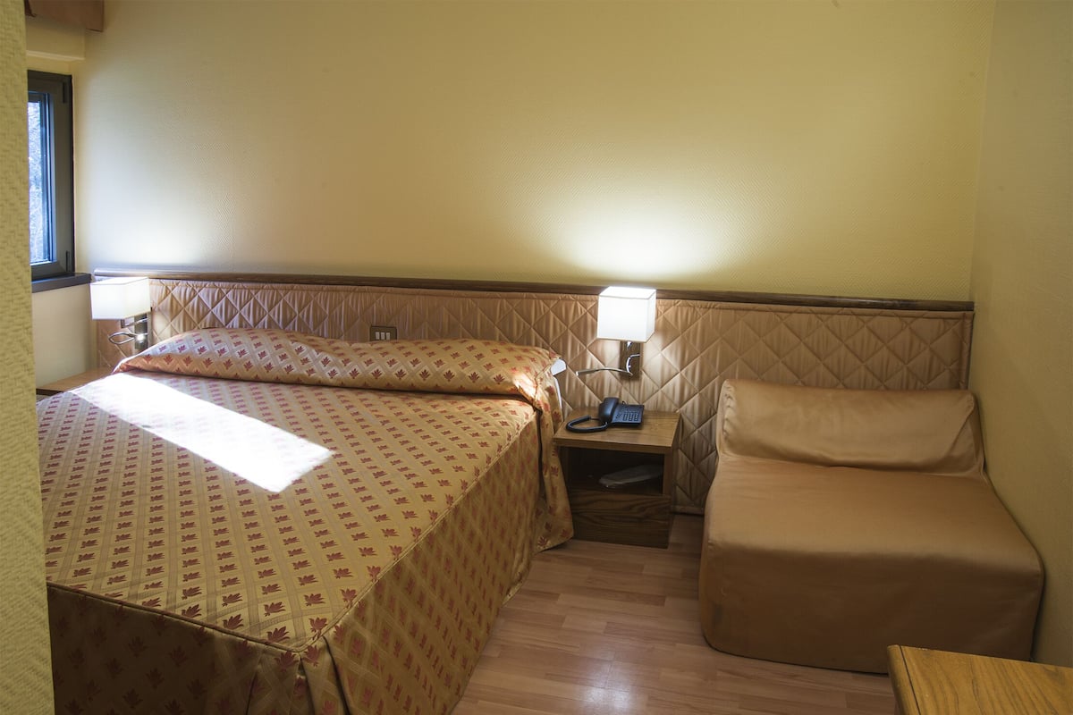 A small, cozy hotel room with a bed, a bedside table with a lamp, and a chair.