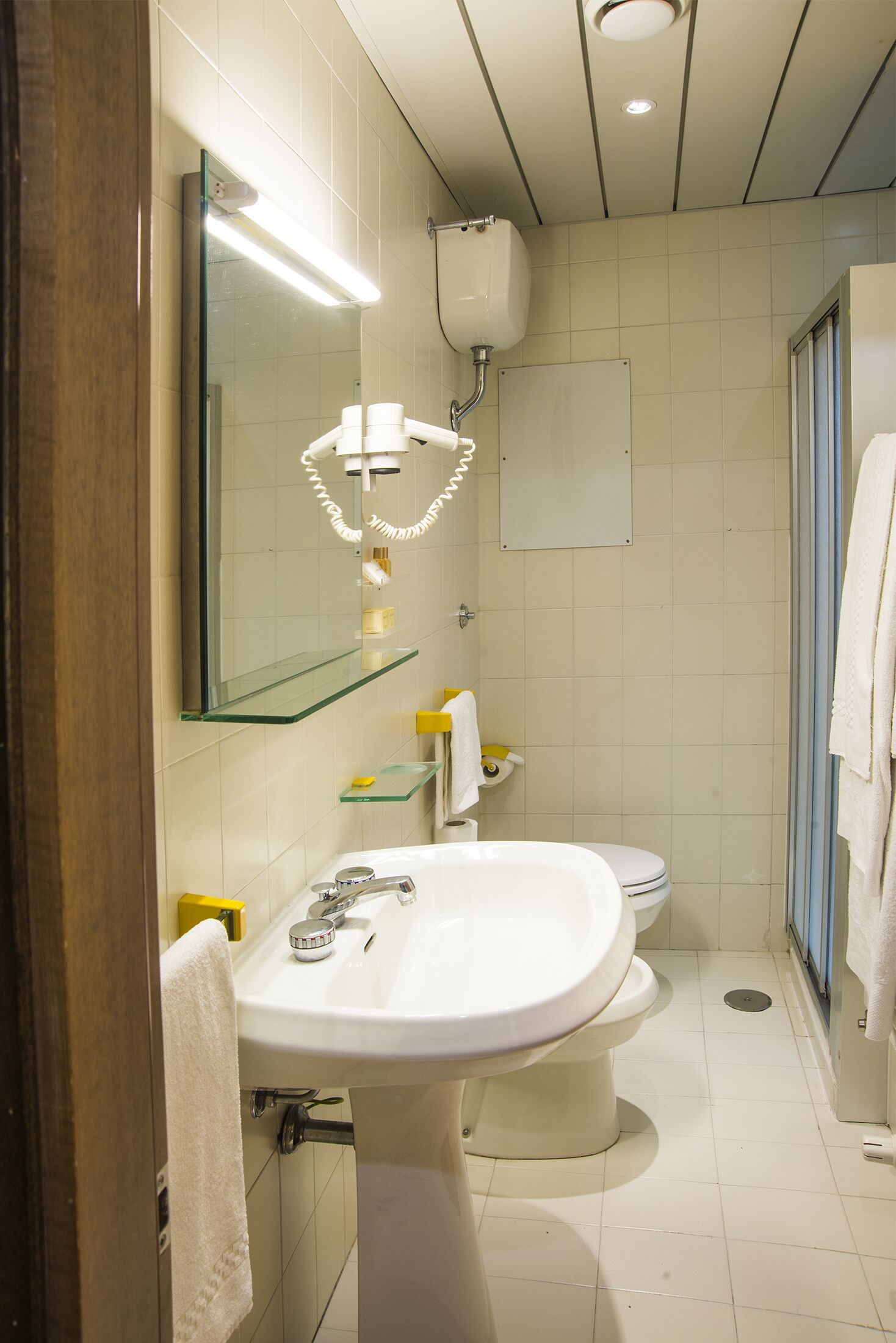 Et badeværelse med hvid vask, spejl og toilet. Der er en glashylde over vasken og et håndklæde hænger på væggen.