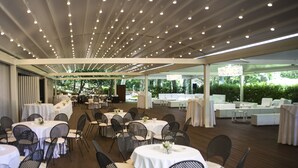 A spacious indoor venue with multiple round tables and chairs, a high ceiling with evenly spaced lights, and a view of greenery outside. - Hotel Giubileo (Pignola)