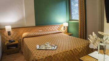Una habitación de hotel con una cama, una mesita de noche con lámpara, una bandeja con tazas de té y una ventana con cortinas.