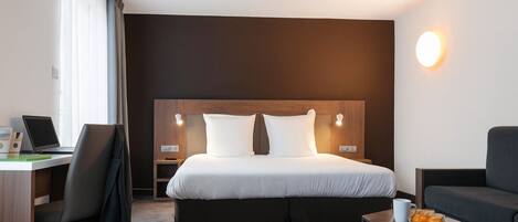 A hotel room with a large bed, a desk with a laptop, a chair, a small table with a cup and oranges, and a nightstand with a lamp.