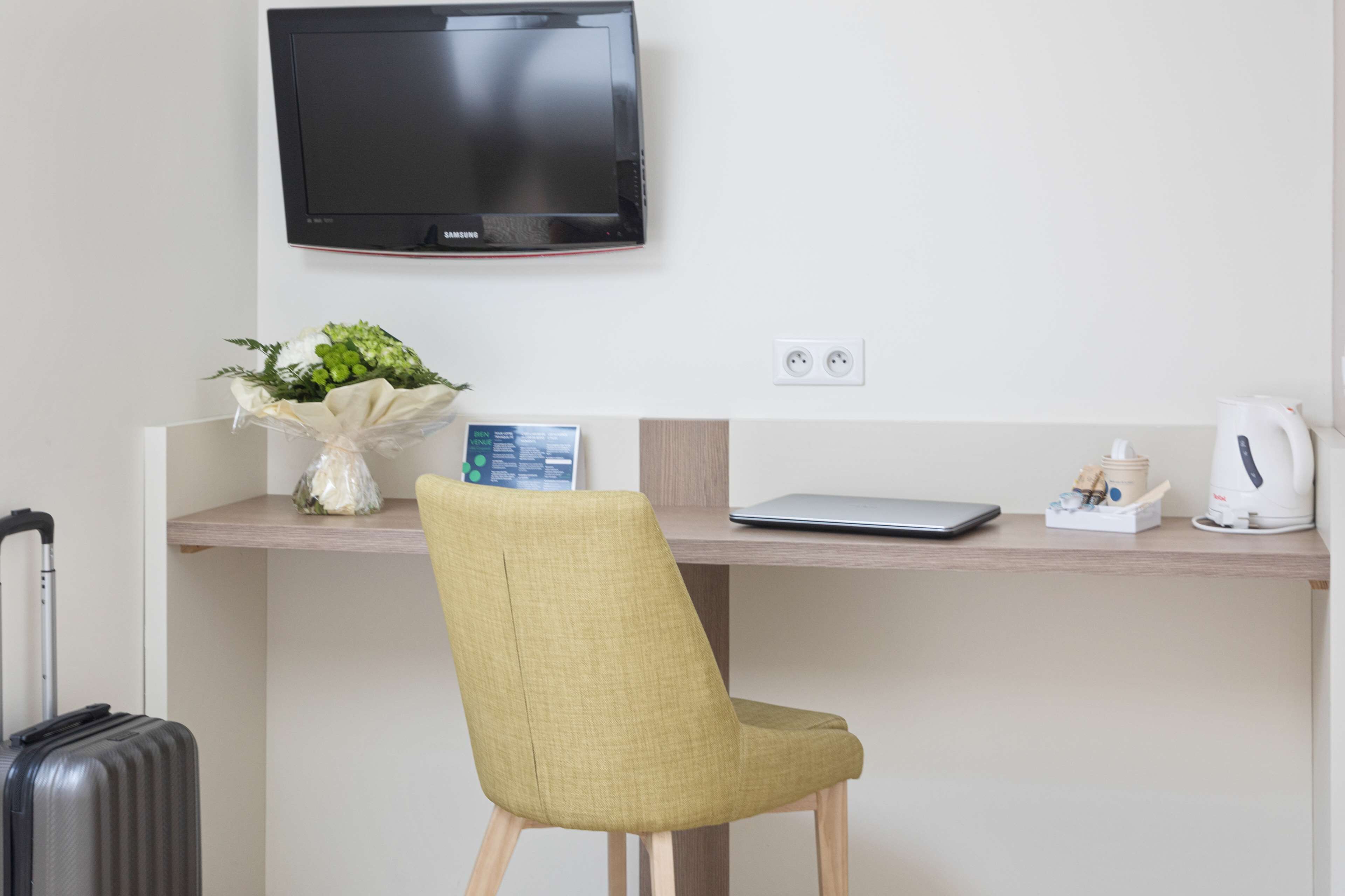 A hotel room with a desk, a chair, a television, a laptop, a coffee maker, and a suitcase.