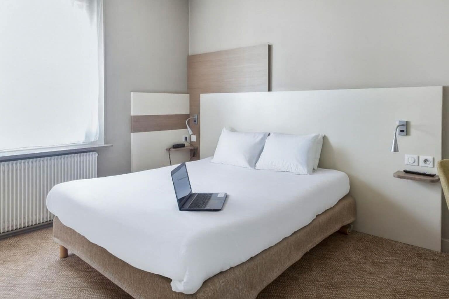 Et moderne hotelværelse med en stor seng, en bærbar computer på sengen, et natbord med en lampe og udsigt over det fri.
