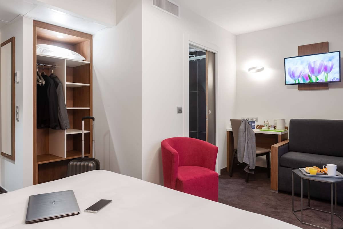 A hotel room with a bed, a desk, a red chair, a TV, and a wardrobe.