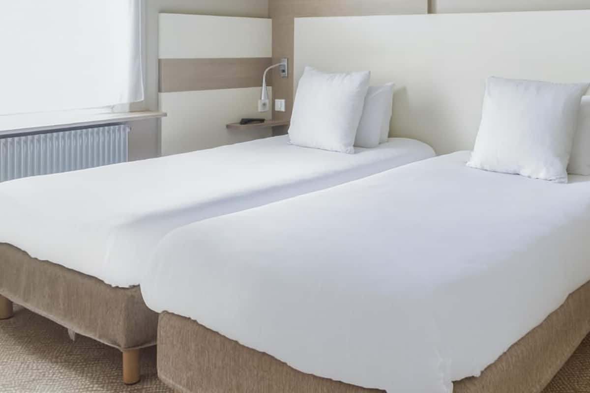 A hotel room with two beds, a bedside table, a lamp, and a window.