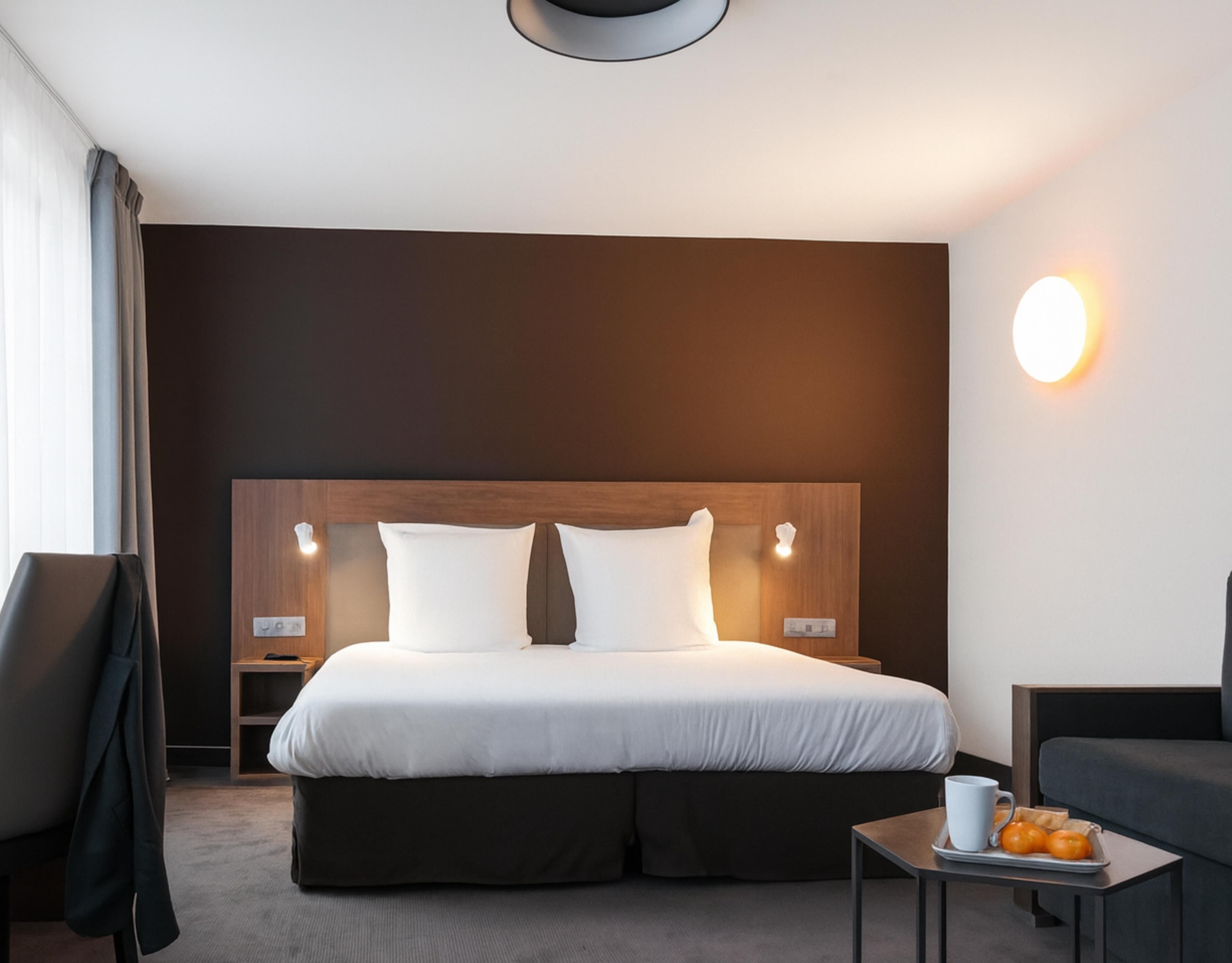 A hotel room with a large bed, bedside tables, a chair, a small table with a tray of oranges, and a wall-mounted light.
