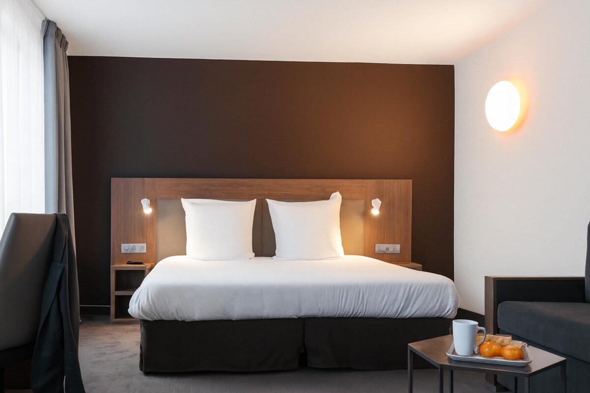 A hotel room with a large bed, bedside tables, a chair, a small table with a tray of oranges, and a wall-mounted light.