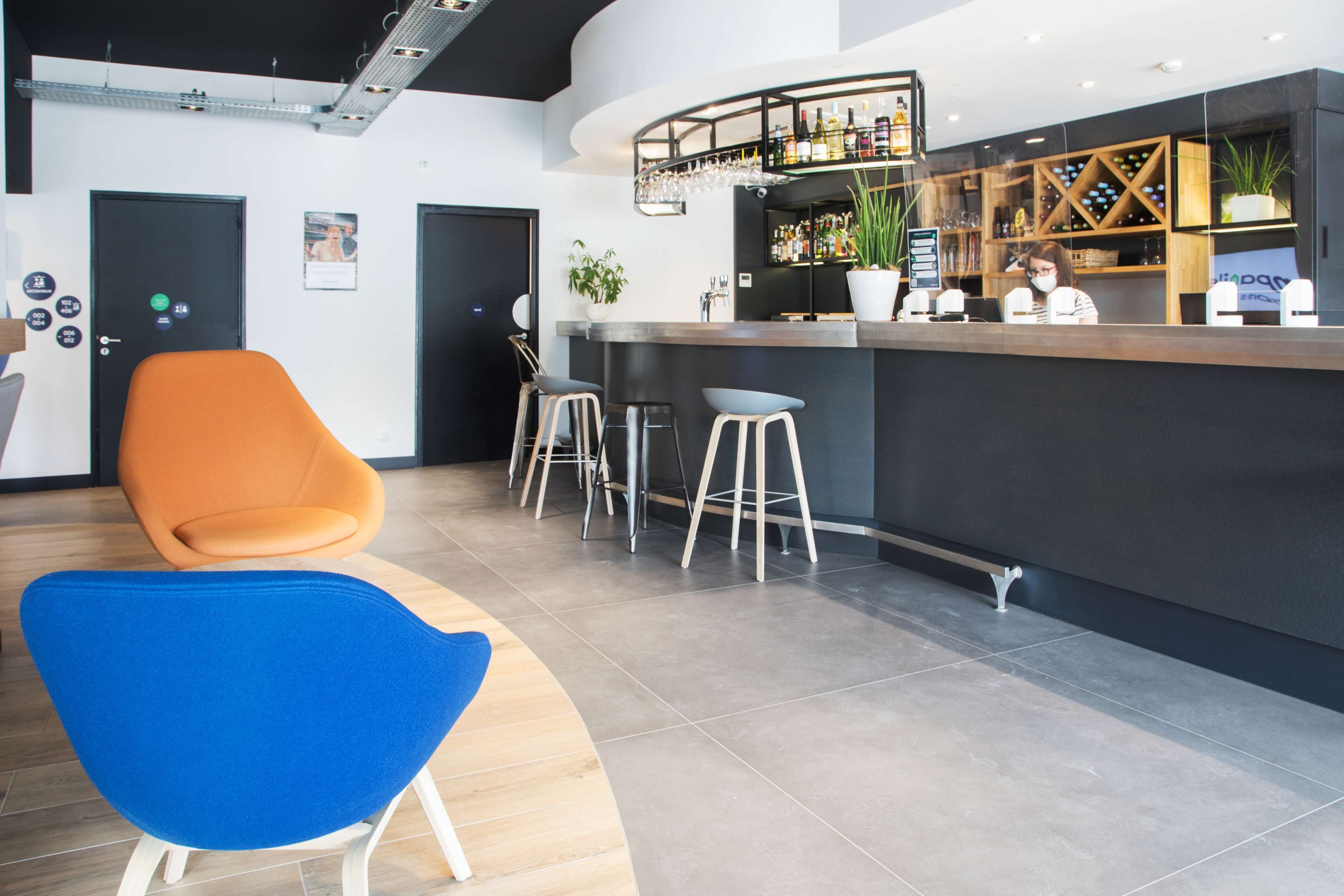 Un espace bar moderne avec un comptoir incurvé, des tabourets de bar et un coin salon comprenant une chaise bleue et une chaise orange.