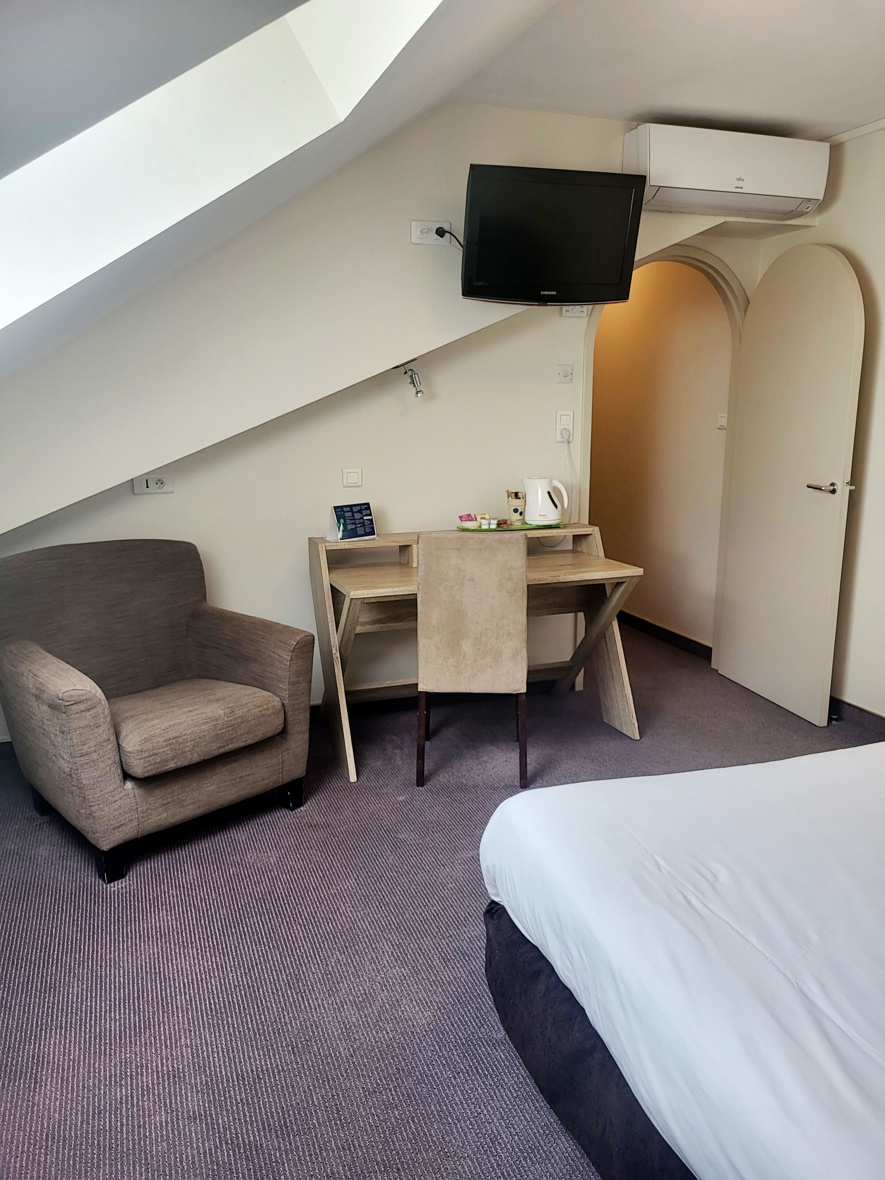 A hotel room with a bed, a desk with a chair, a sofa, a TV mounted on the wall, and an air conditioning unit.