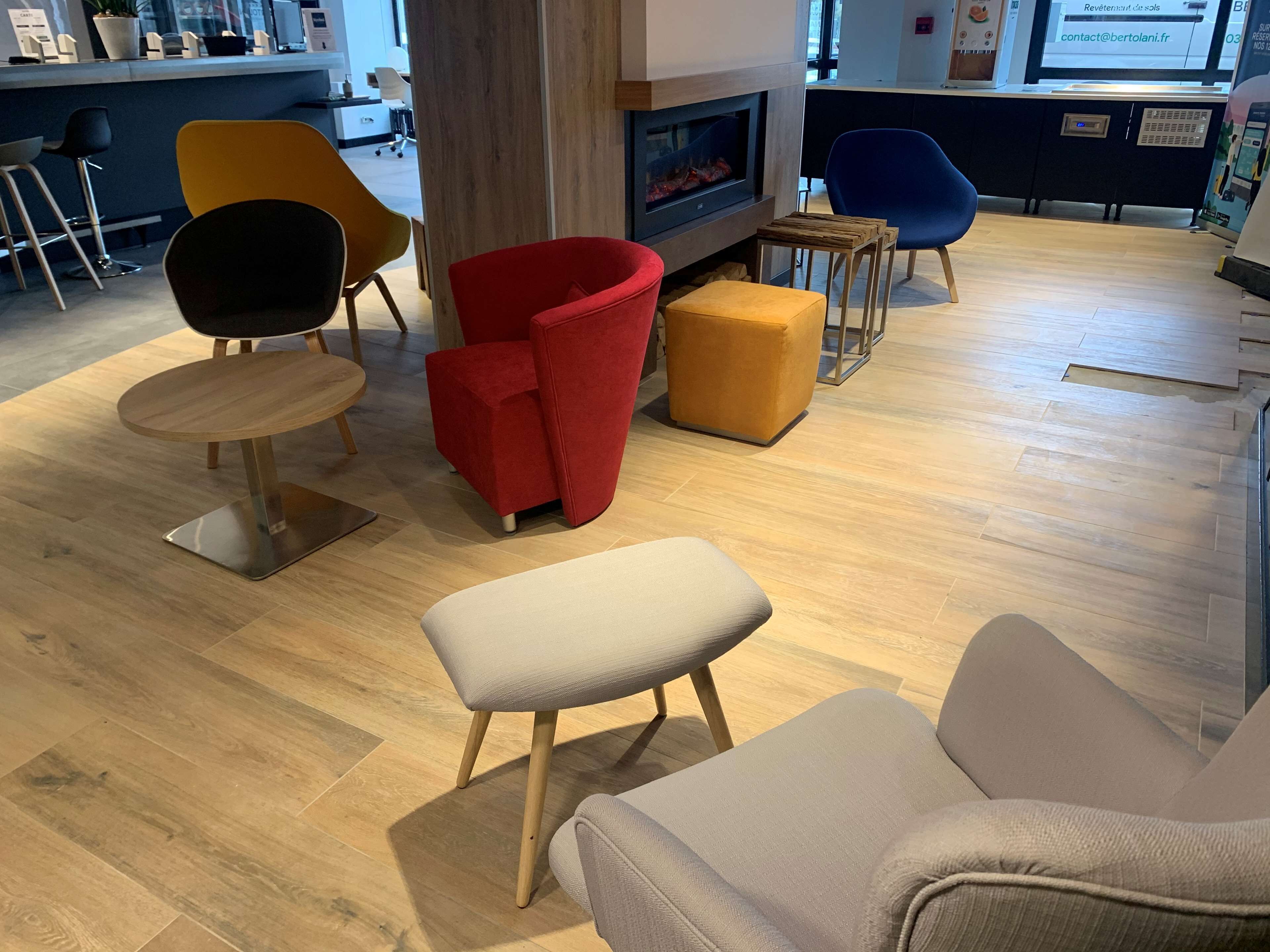 A modern interior with a wooden floor, a fireplace, and various seating options including a red armchair, a yellow chair, and a white ottoman.