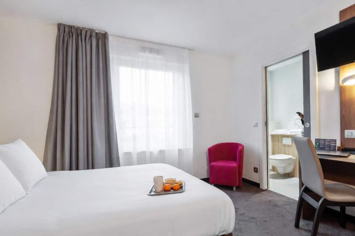 A hotel room with a bed, a desk, a chair, a bathroom, and a window with curtains.