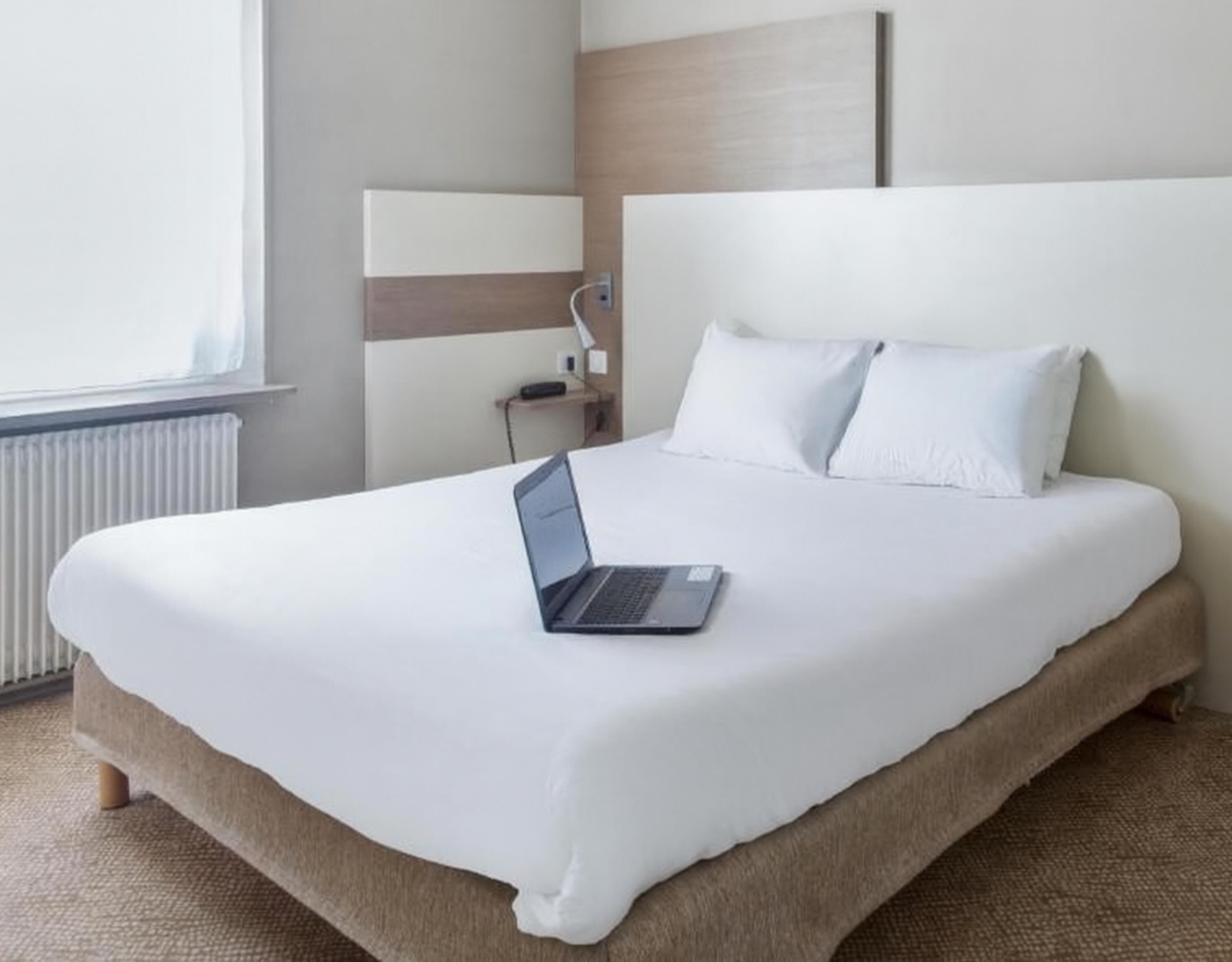 Una cama bien hecha con un ordenador portátil encima, una mesita de noche y una ventana con persianas.