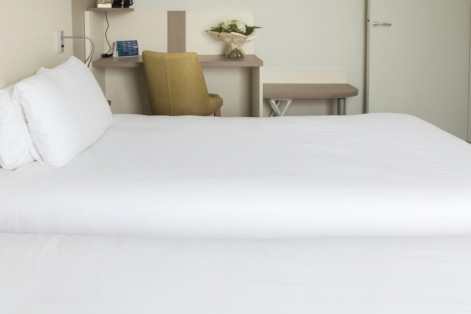 A hotel room with a large bed, a desk with a chair, a small table, and a wall-mounted shelf with a vase of flowers.
