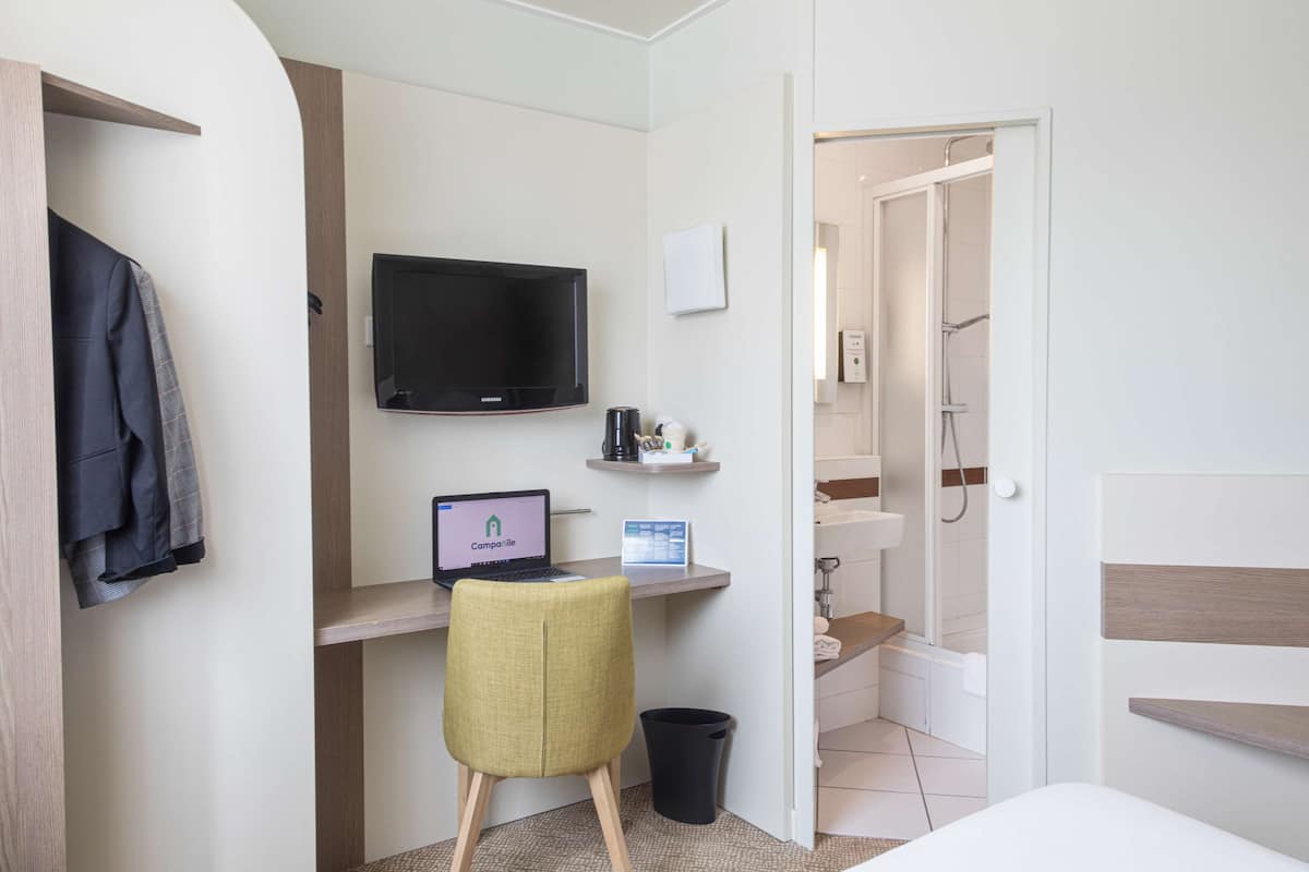 A hotel room with a desk, a television, a laptop, a chair, a small trash bin, and a shower area.