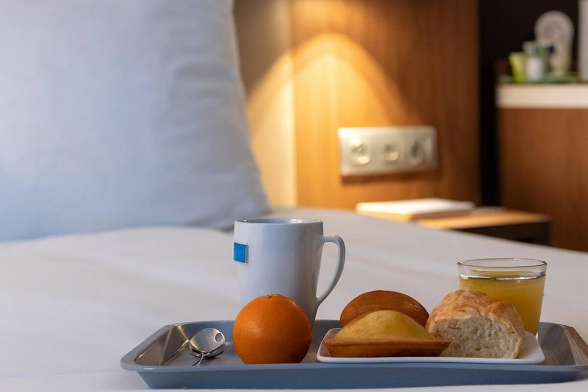 A tray with a mug, orange, and pastries on a bed with a pillow and a nightstand with a lamp.