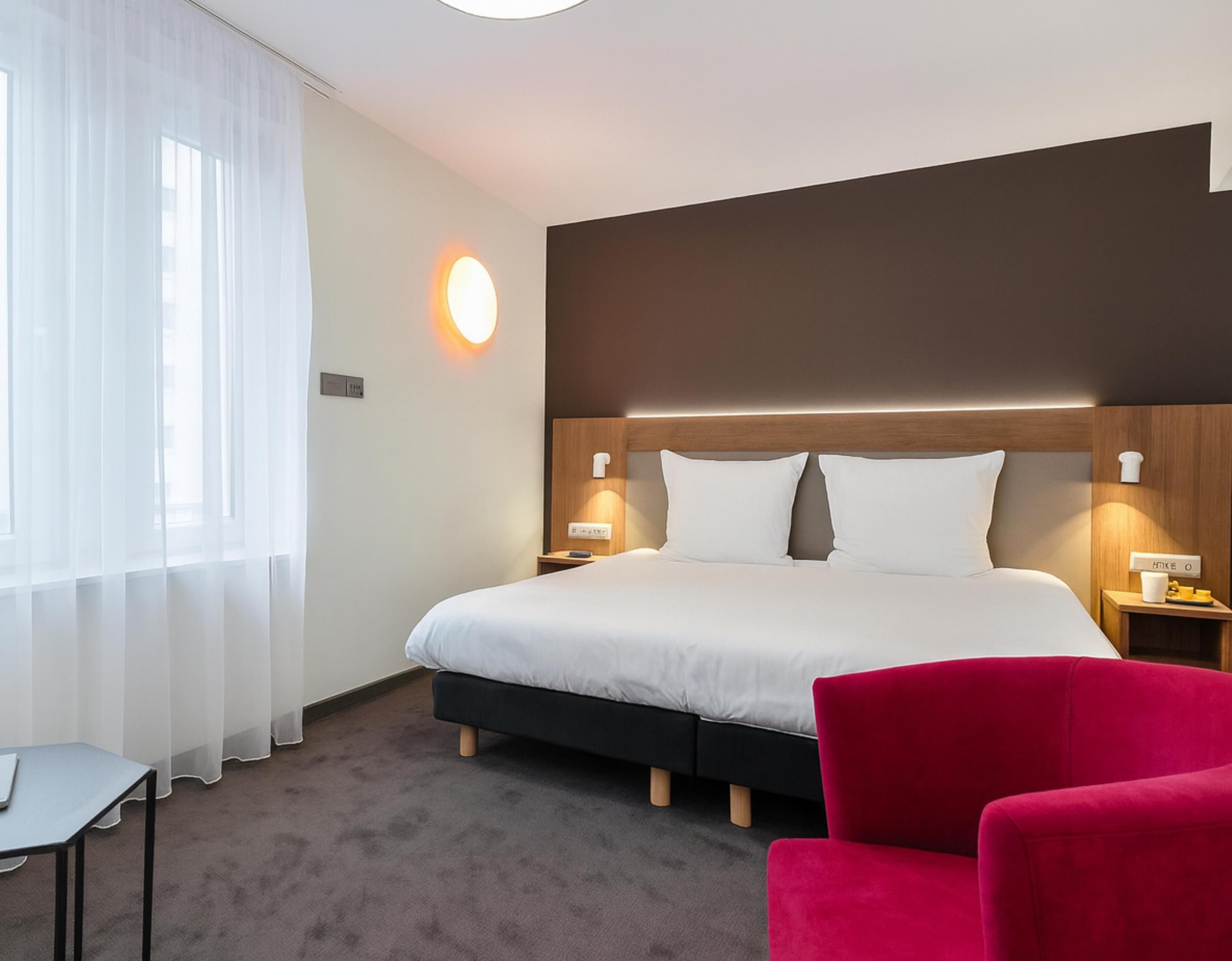 A hotel room with a large bed, a red chair, a desk, and a window with sheer curtains.