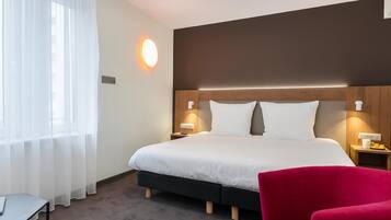 A hotel room with a large bed, a red chair, a desk, and a window with sheer curtains.