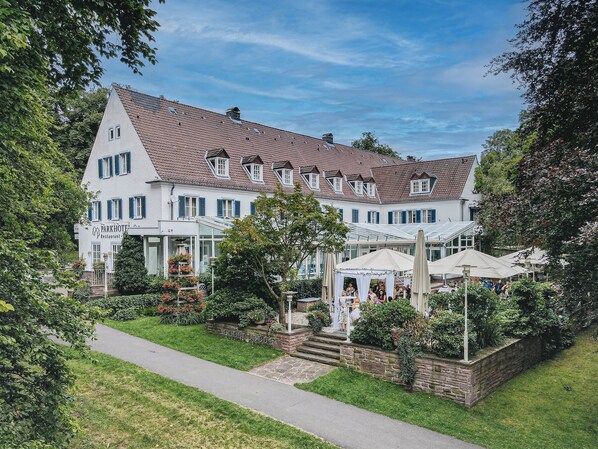 Ein großes, weißes Gebäude mit einem Ziegeldach und zahlreichen Fenstern, umgeben von üppiger Vegetation und einem gepflasterten Weg.