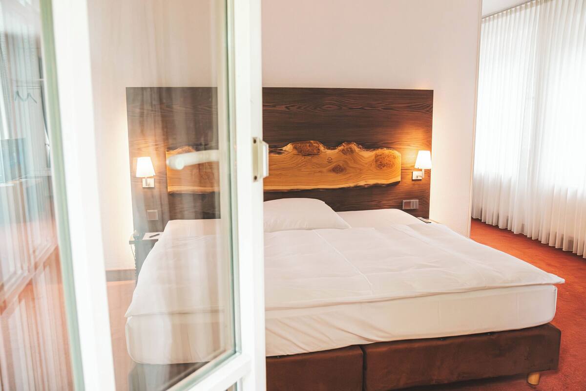 A hotel room with a wooden headboard, a bed with white linens, two bedside lamps, and a view through a glass door.