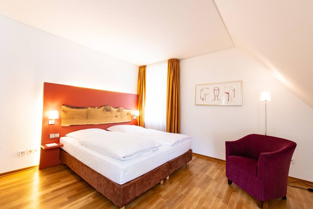 A hotel room with a bed, a red headboard, a red armchair, a wall-mounted lamp, and a framed picture.