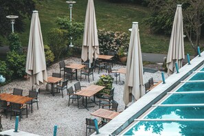 Eine Terrasse im Freien mit Tischen und Stühlen, umgeben von Sonnenschirmen, und ein Schwimmbecken im Hintergrund.