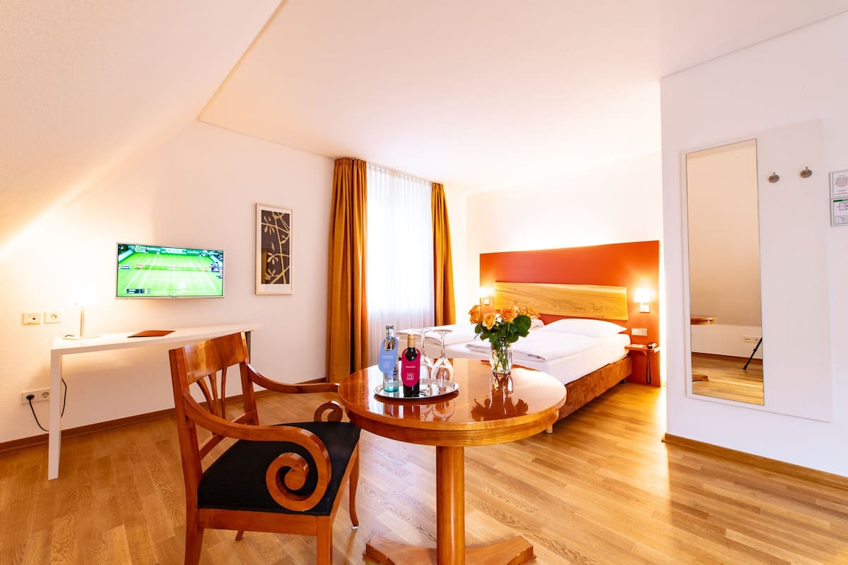 A hotel room with a bed, a desk with a TV, a dining table with chairs, and a small table with a vase of flowers.