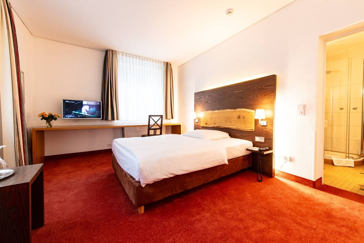 A hotel room with a large bed, a desk with a TV, a shower, and a red carpet.