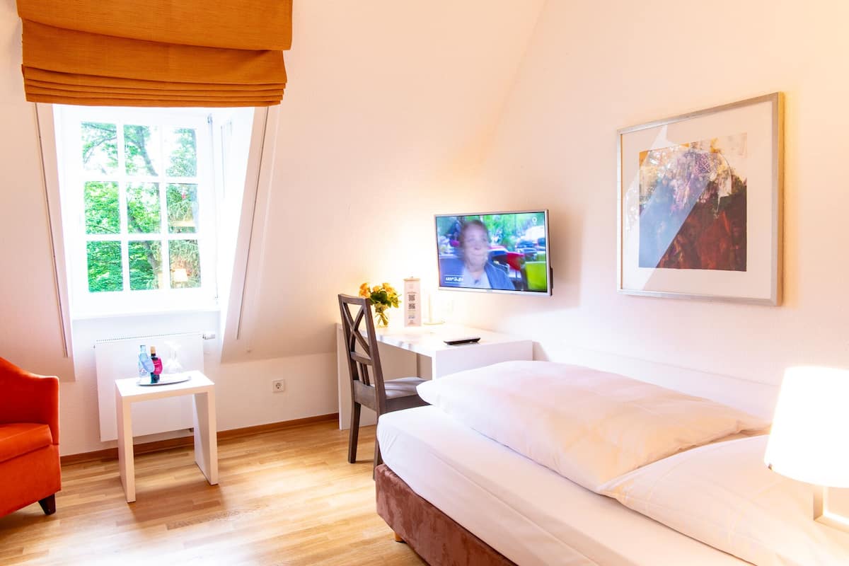 A hotel room with a bed, a desk with a television, a chair, a small table with a bottle, and a window with curtains.