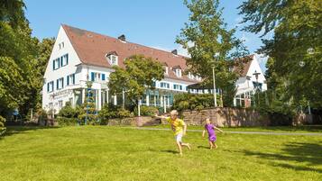 Traditional European-style hotel with a white facade and red-tiled roof, set amidst a lush, grassy area with mature trees.