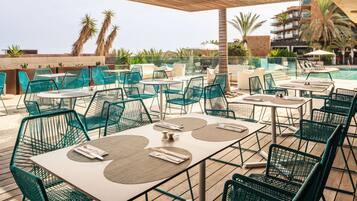 Un espace extérieur aménagé pour les repas, avec des tables et des chaises, une piscine en arrière-plan et des palmiers.