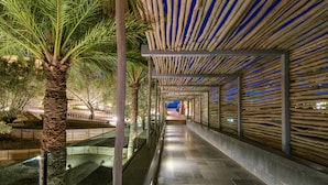 A covered walkway with a glass barrier, palm trees, and a wooden ceiling structure. - Salobre Hotel Resort & Serenity (San Bartolomé de Tirajana)
