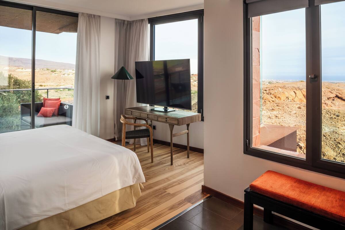 Et hotelværelse med en stor seng, et skrivebord med tv, en stol, en bænk og udsigt til ørkenlandskabet.