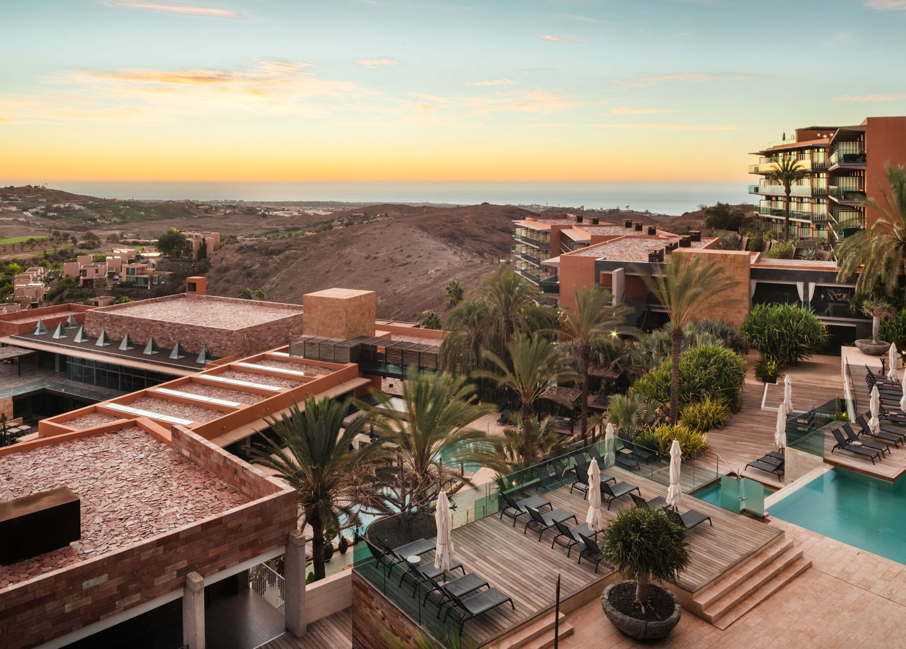 A resort with a rooftop pool, lounge areas, and a view of the ocean at sunset.