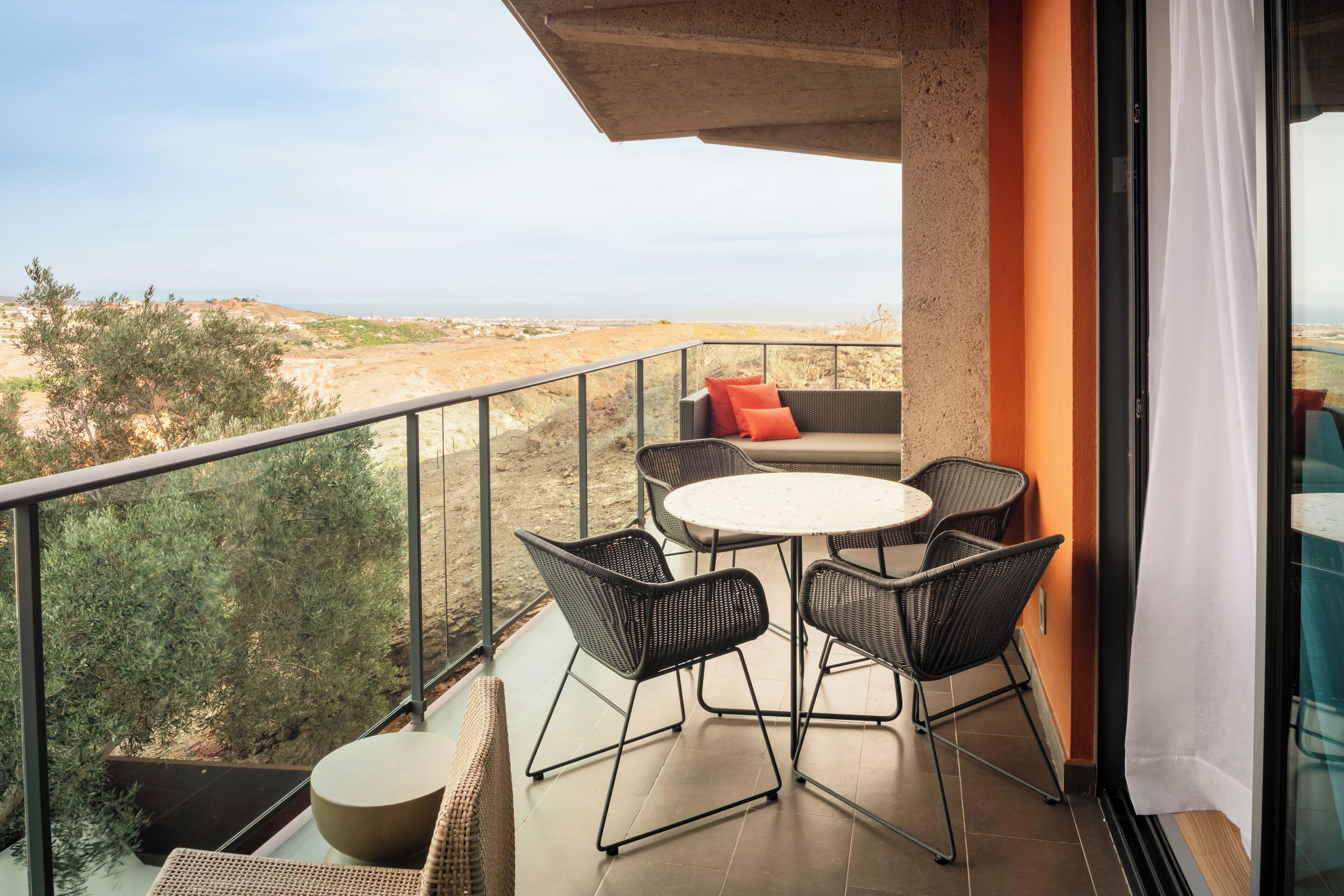 Een balkon met een ronde tafel en vier stoelen, een bank met een rode kussen, en uitzicht op het landschap.